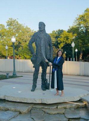 Graduate in cap and gown poses beside a large bronze statue on a stone platform outdoors.