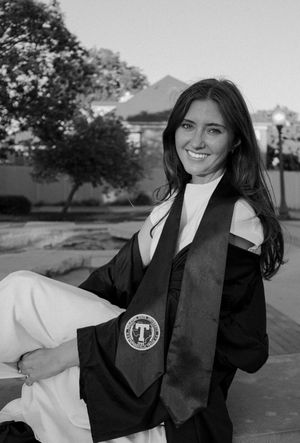 Smiling young woman in black graduation gown and stole poses outdoors in black and white photo.