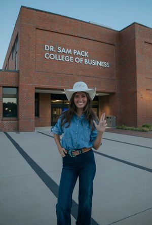Young woman in cowboy hat and denim outfit standing in front of Dr. Sam Pack College of Business building.