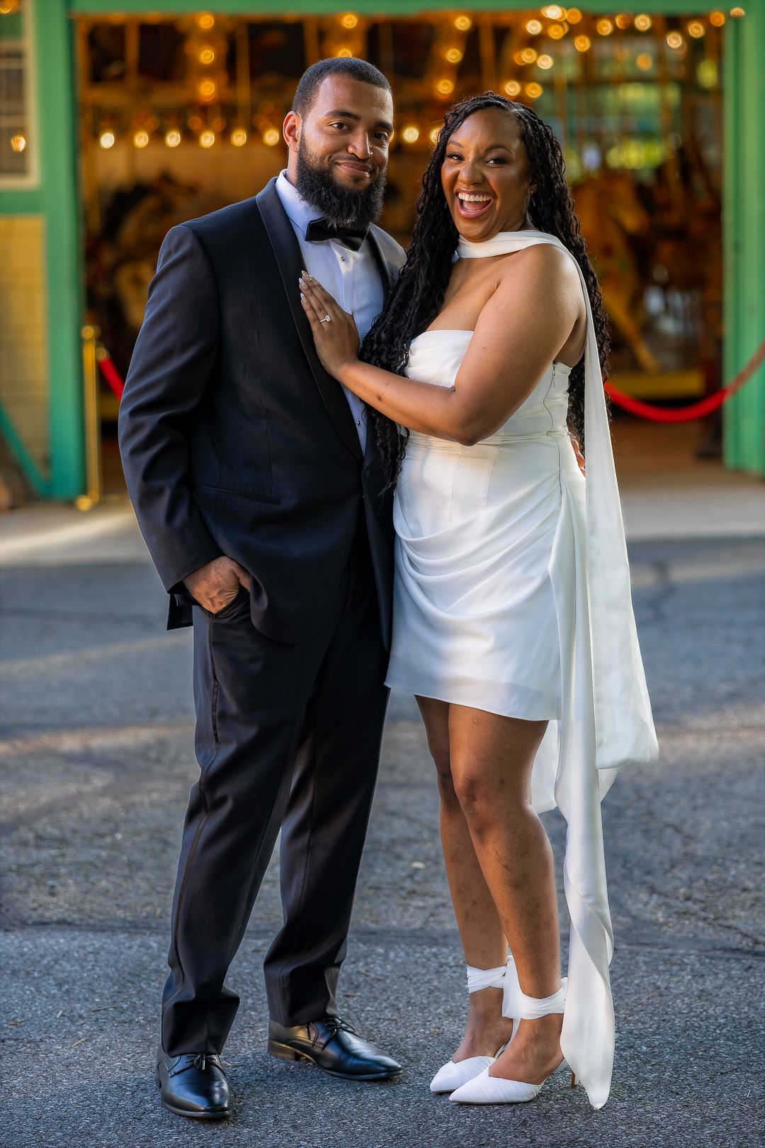 Elegant couple posing in formal attire, man in black tuxedo and woman in white mini dress, smiling in front of a carousel.