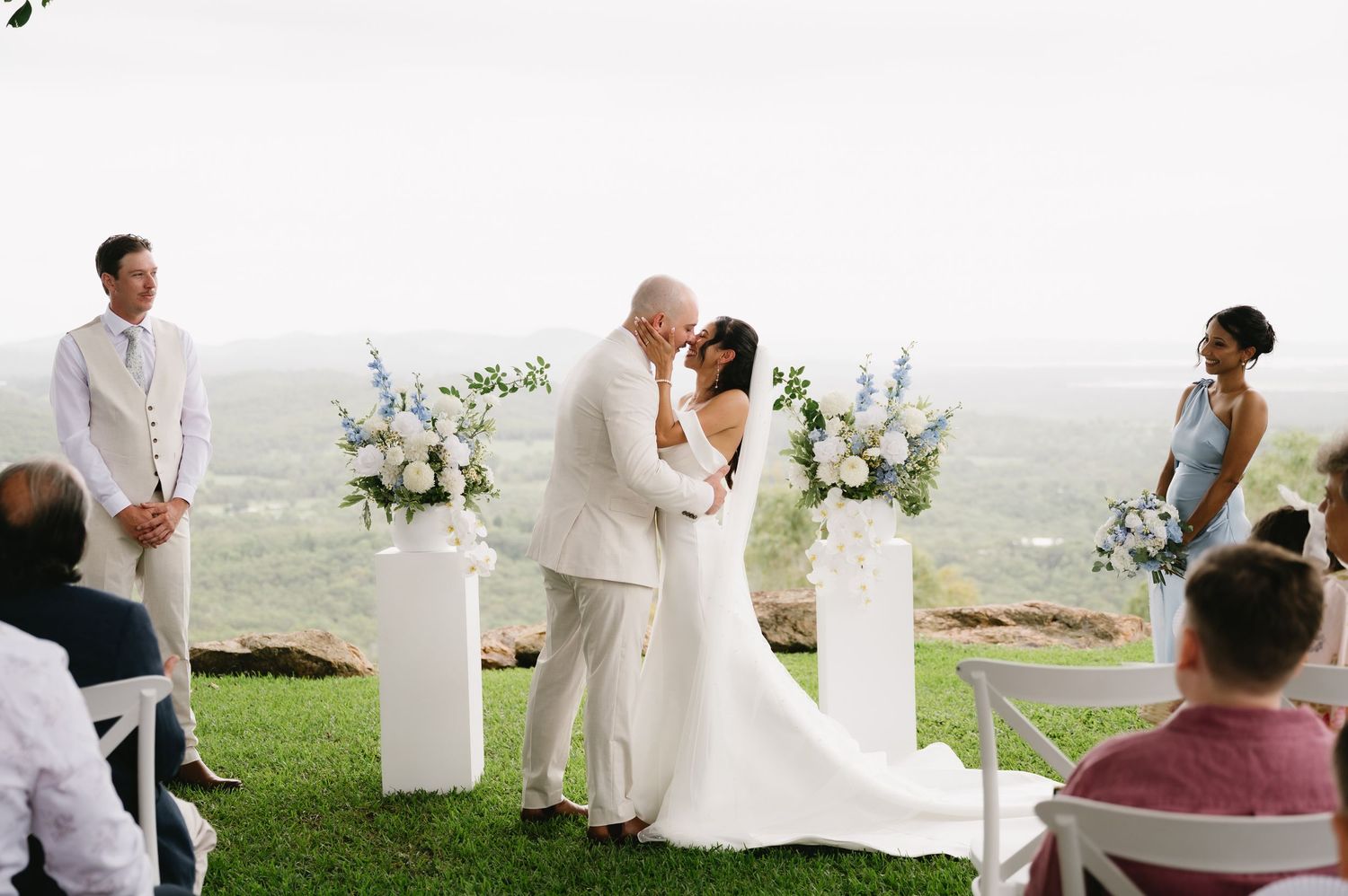 Bride and groom share first kiss at outdoor wedding ceremony with white floral arrangements and scenic valley views.