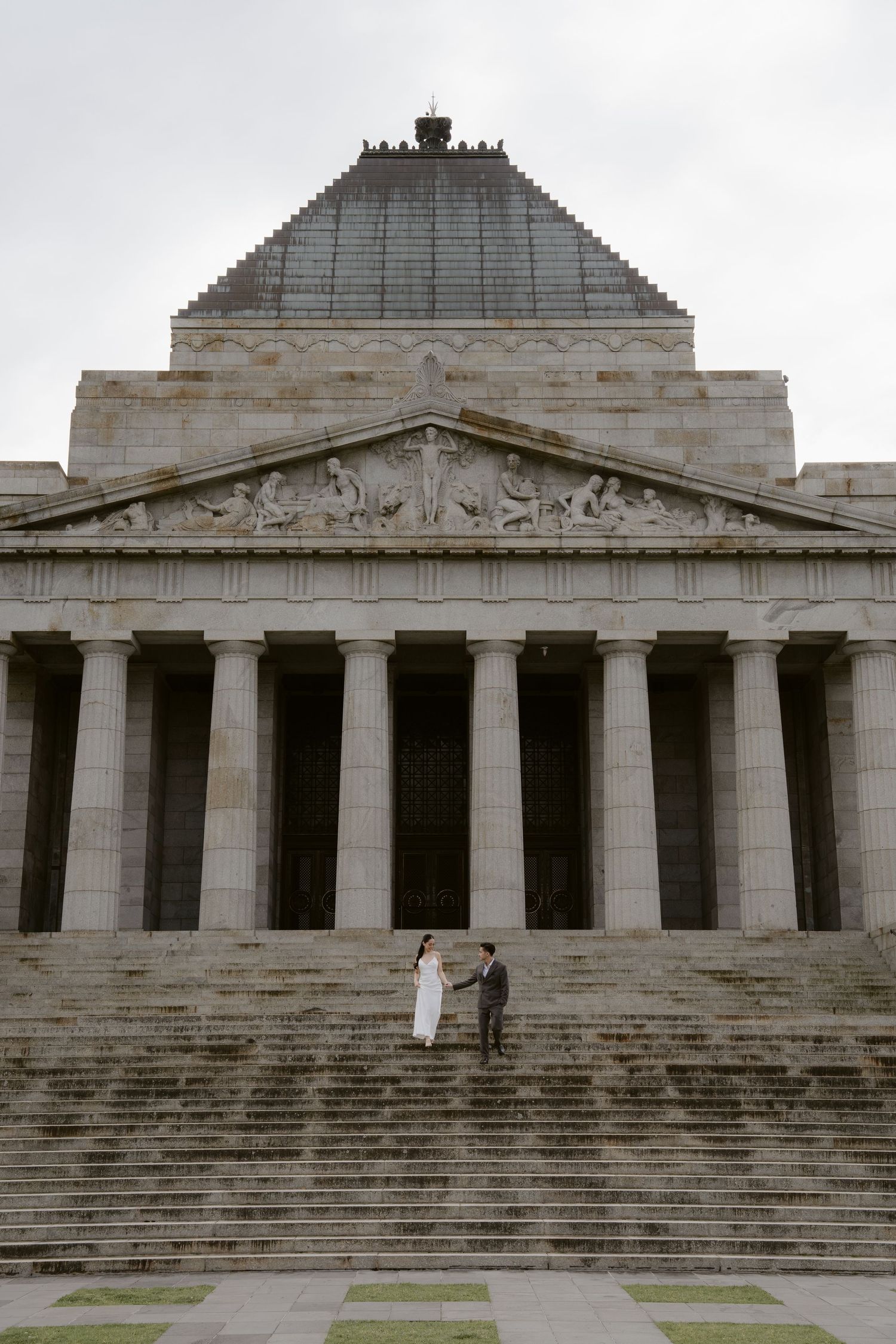 Couple in wedding attire stands on steps of the Shrine of Remembrance in Melbourne, Australia.