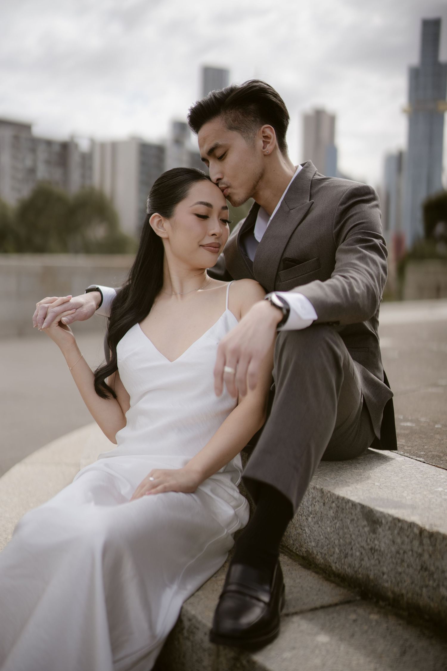 Couple in wedding attire sitting on steps with city skyline, groom in gray suit kissing bride in white dress.