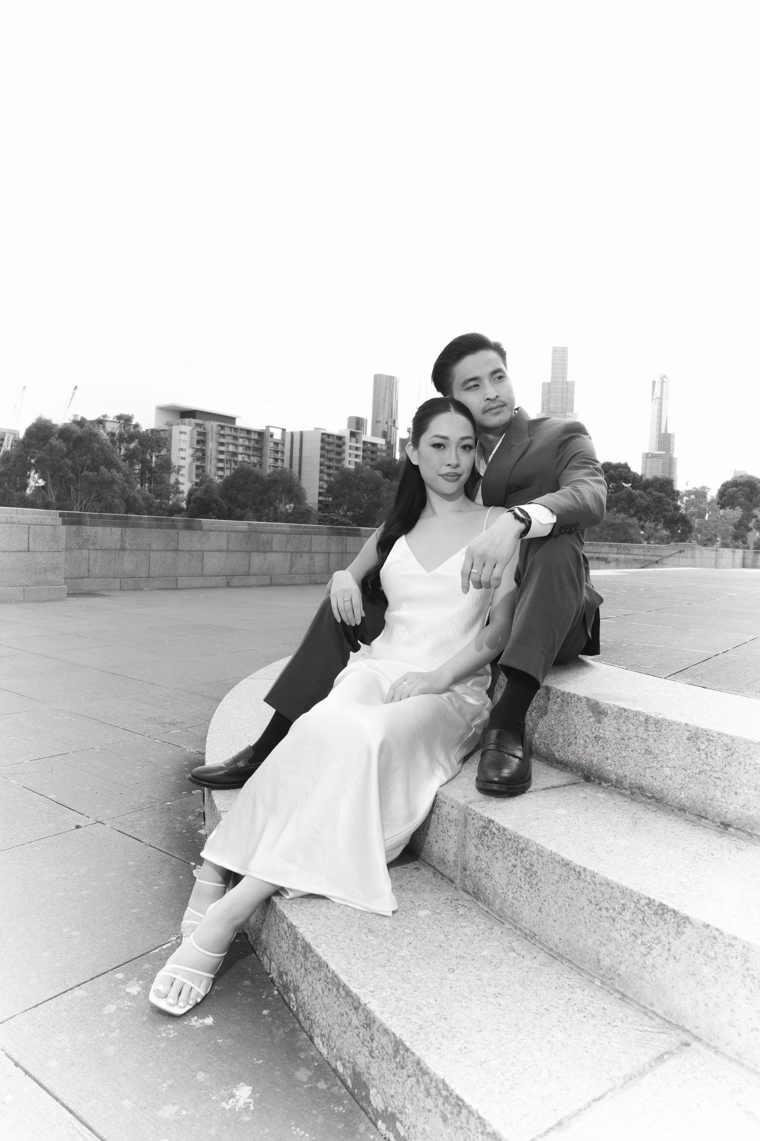 Black and white photo of a couple in formal wear sitting on outdoor steps with a city skyline in the background.