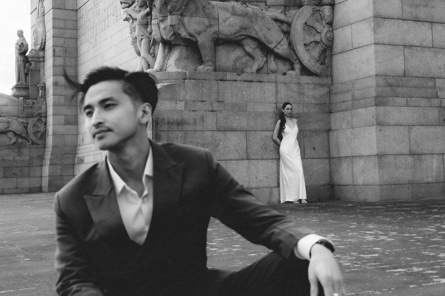 Black and white photo of a young man in a suit sitting near ornate stone columns with a woman in white dress standing behind.