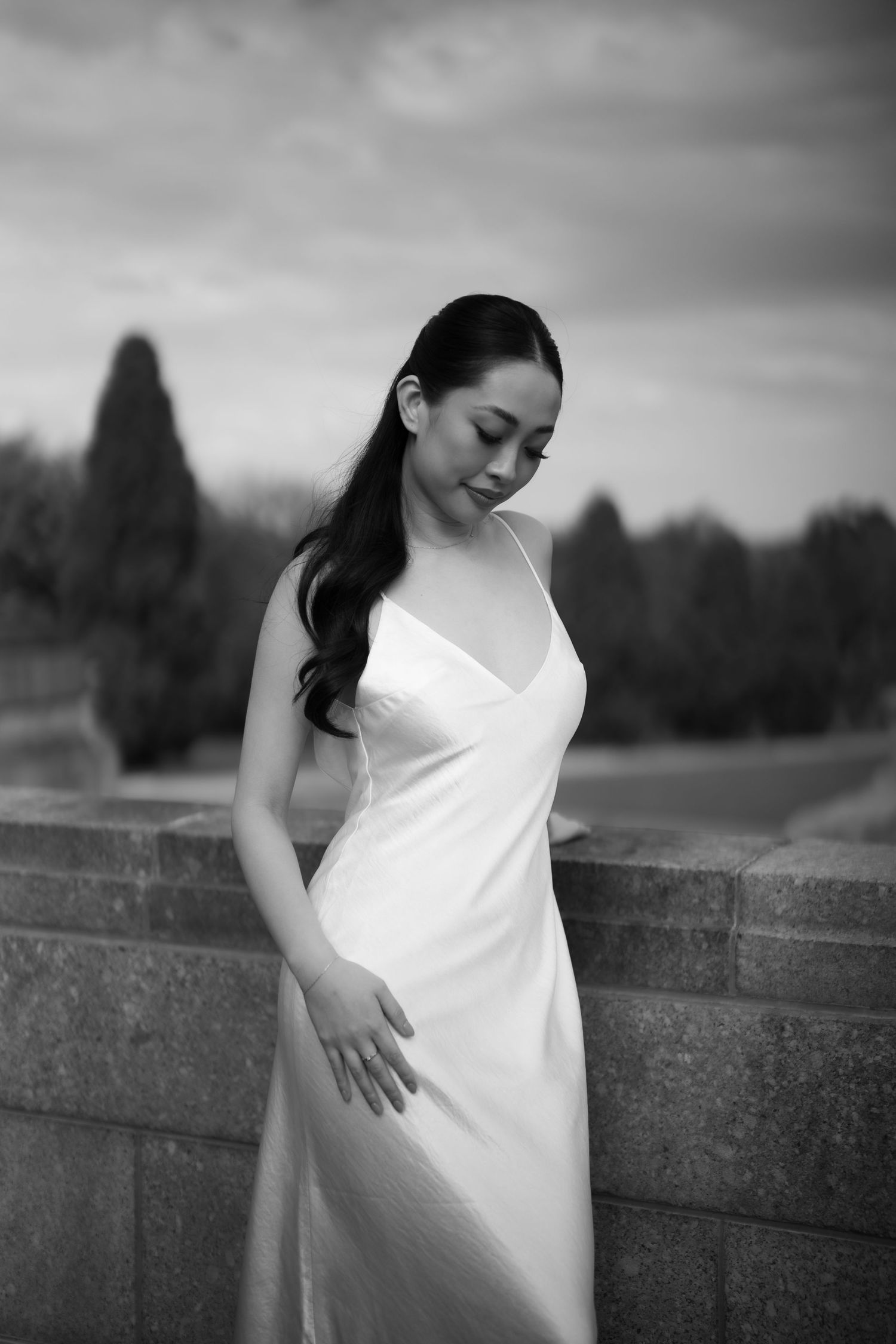 Black and white portrait of a woman in a white slip dress posing gracefully outdoors near a stone wall.
