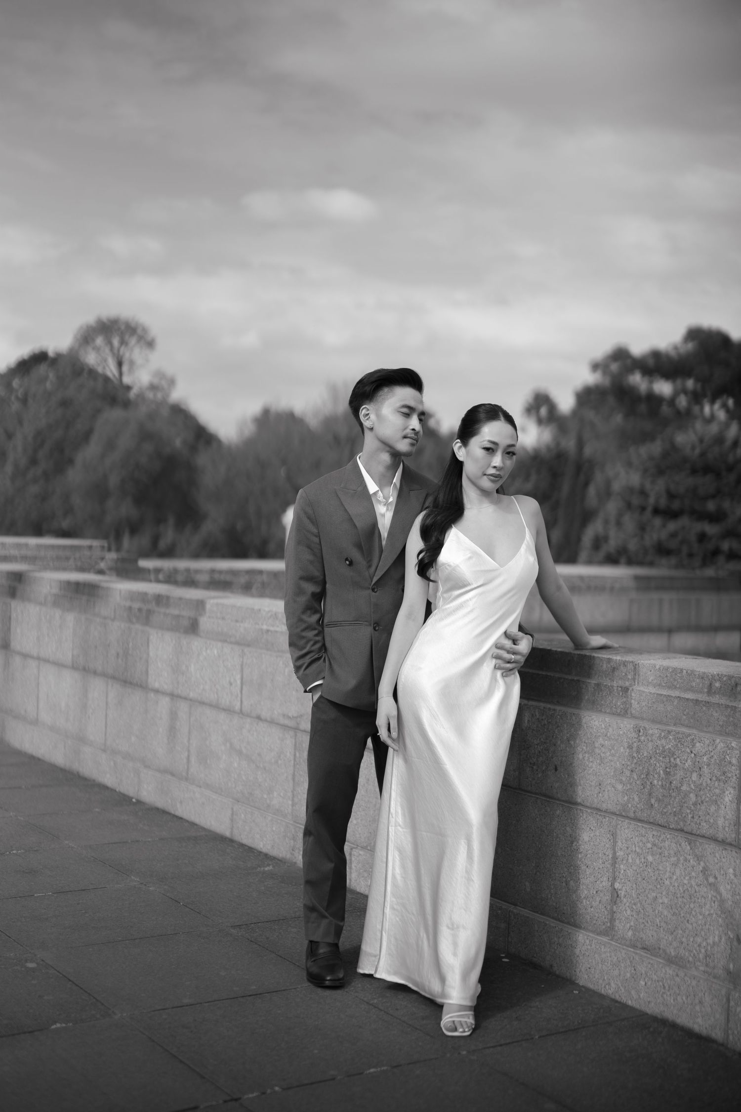 Black and white photo of an elegant couple in wedding attire posing by a stone wall with trees in the background.