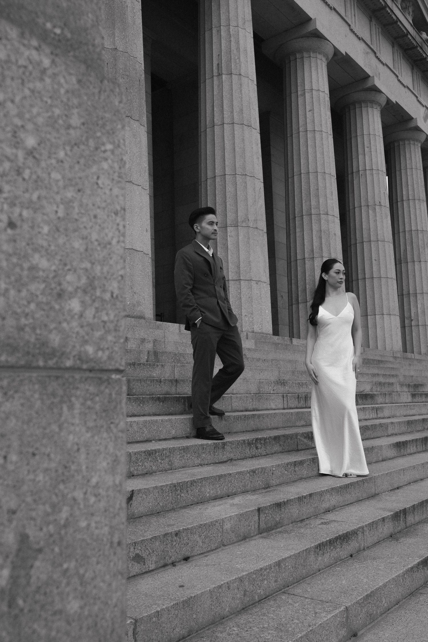 Couple in wedding attire pose on grand stone steps with classical columns in black and white.
