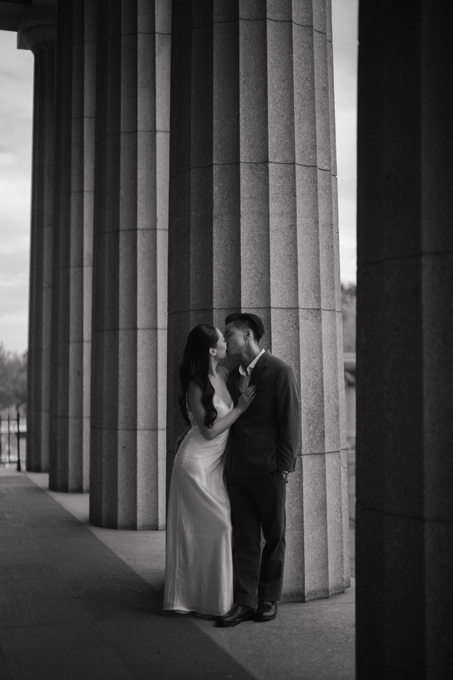 Black and white photo of a couple in formal wear sharing a romantic moment between large stone columns.