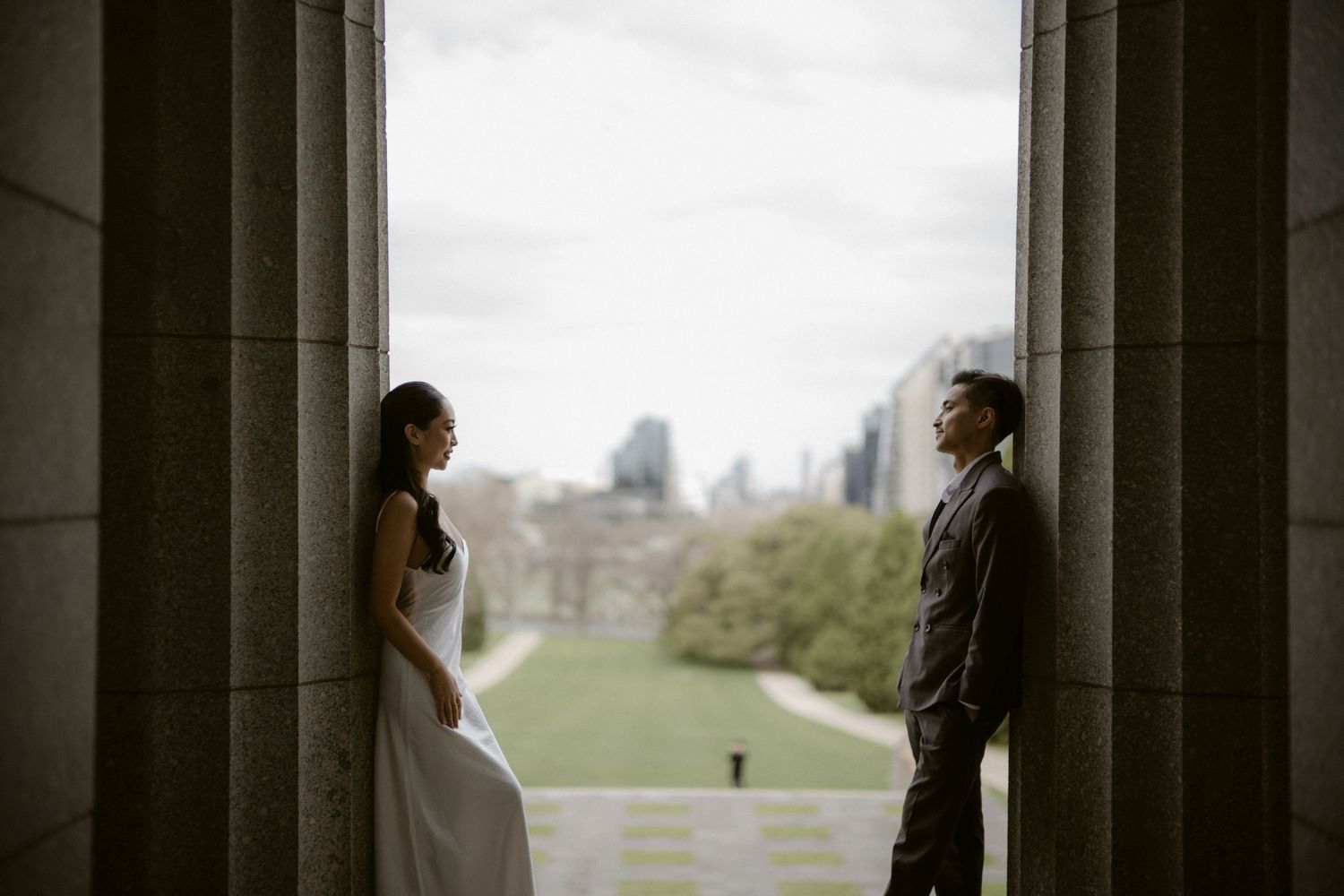 Bride and groom stand facing each other between stone columns with a city skyline and green lawn visible behind them.