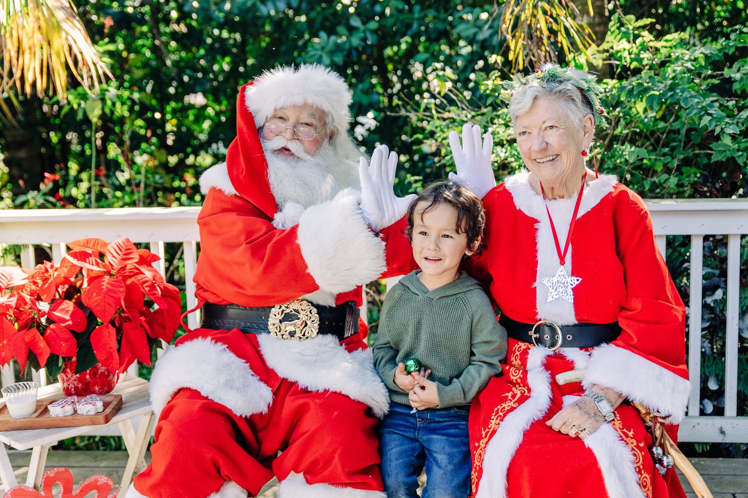 Lake Worth Beach Santa photos of a smiling boy with Santa and Mrs. Claus surrounded by festive poinsettias.