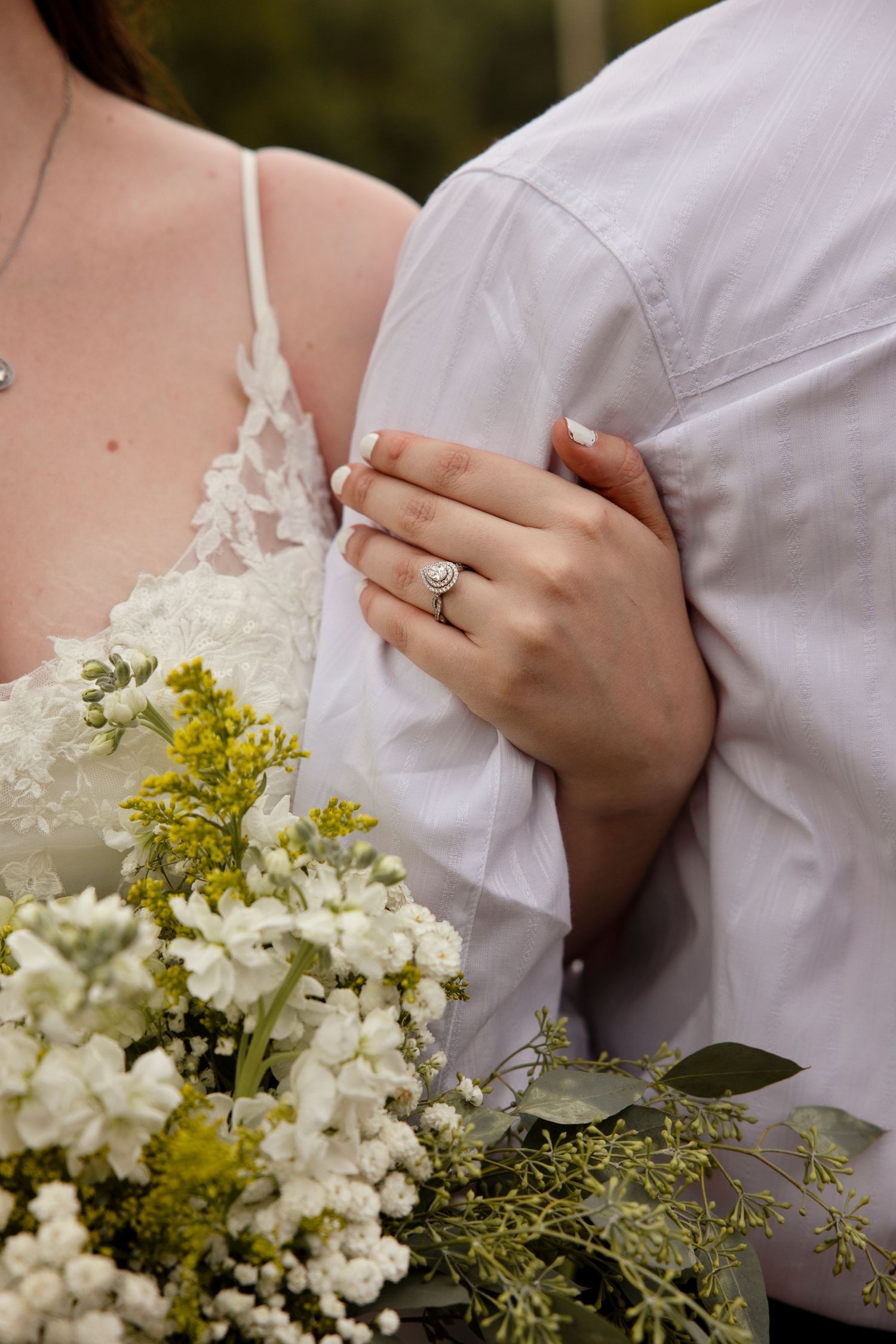 Bride holding white and yellow wildflower bouquet, showcasing halo diamond engagement ring while embracing groom in white shirt.