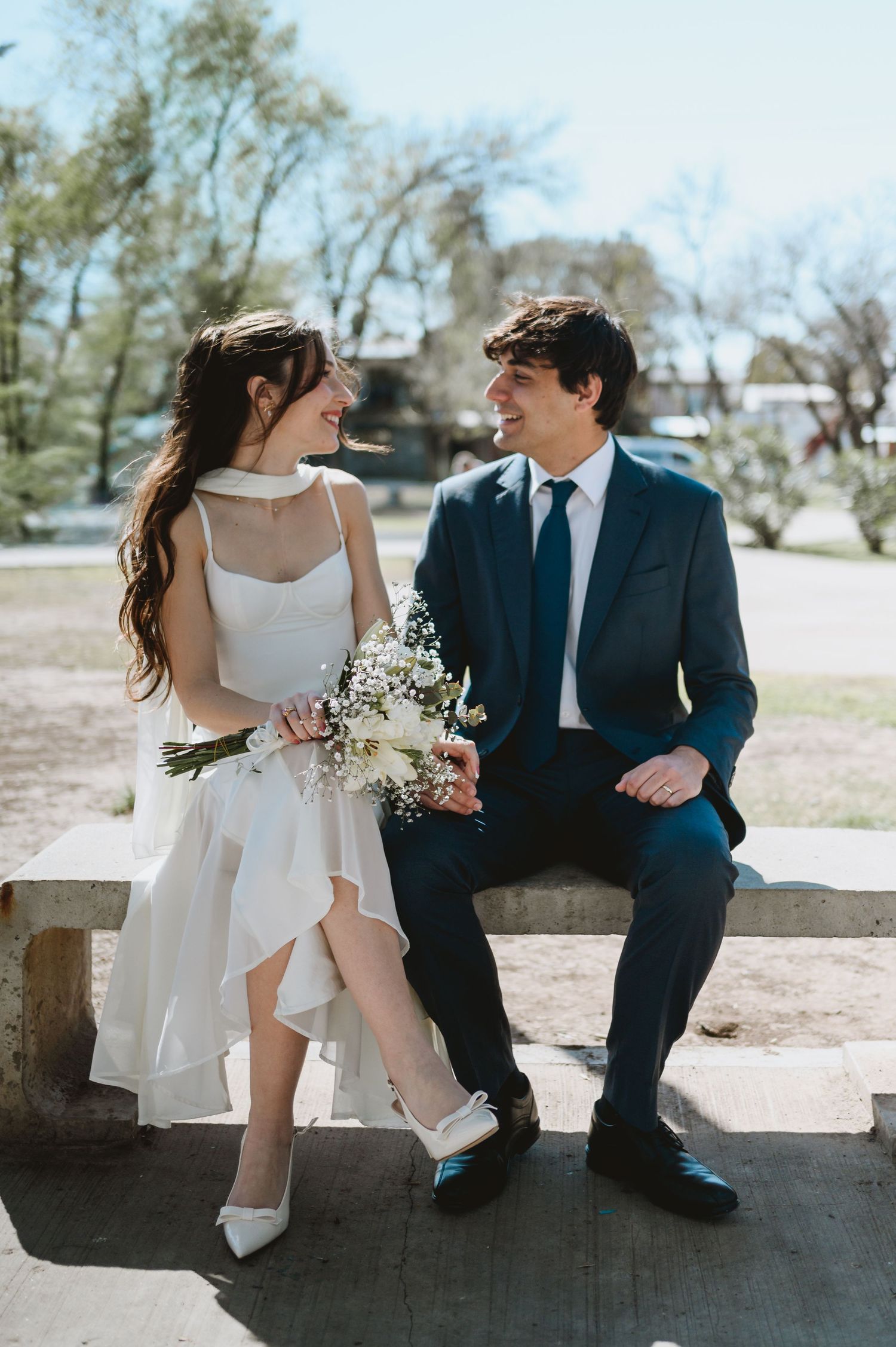A bride and groom sit on a stone bench outdoors, smiling at each other, she holds a wildflower bouquet.