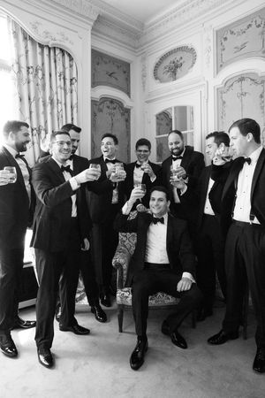 Groom and groomsmen in tuxedos toasting with drinks in an elegant room, black and white wedding photo.