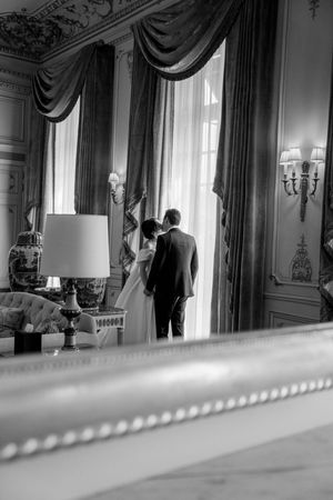 Black and white photo of a bride and groom sharing a kiss by elegant draped windows in a grand ornate room.