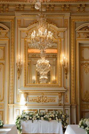 Elegant ballroom with gold ornate moldings, crystal chandelier, mirror, and floral arrangements on a white draped table.
