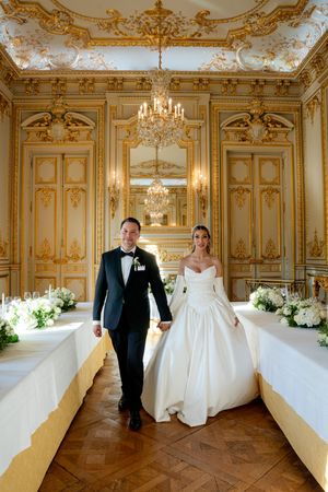Bride and groom walk hand in hand through an ornate gold Parisian ballroom with chandelier and floral arrangements.