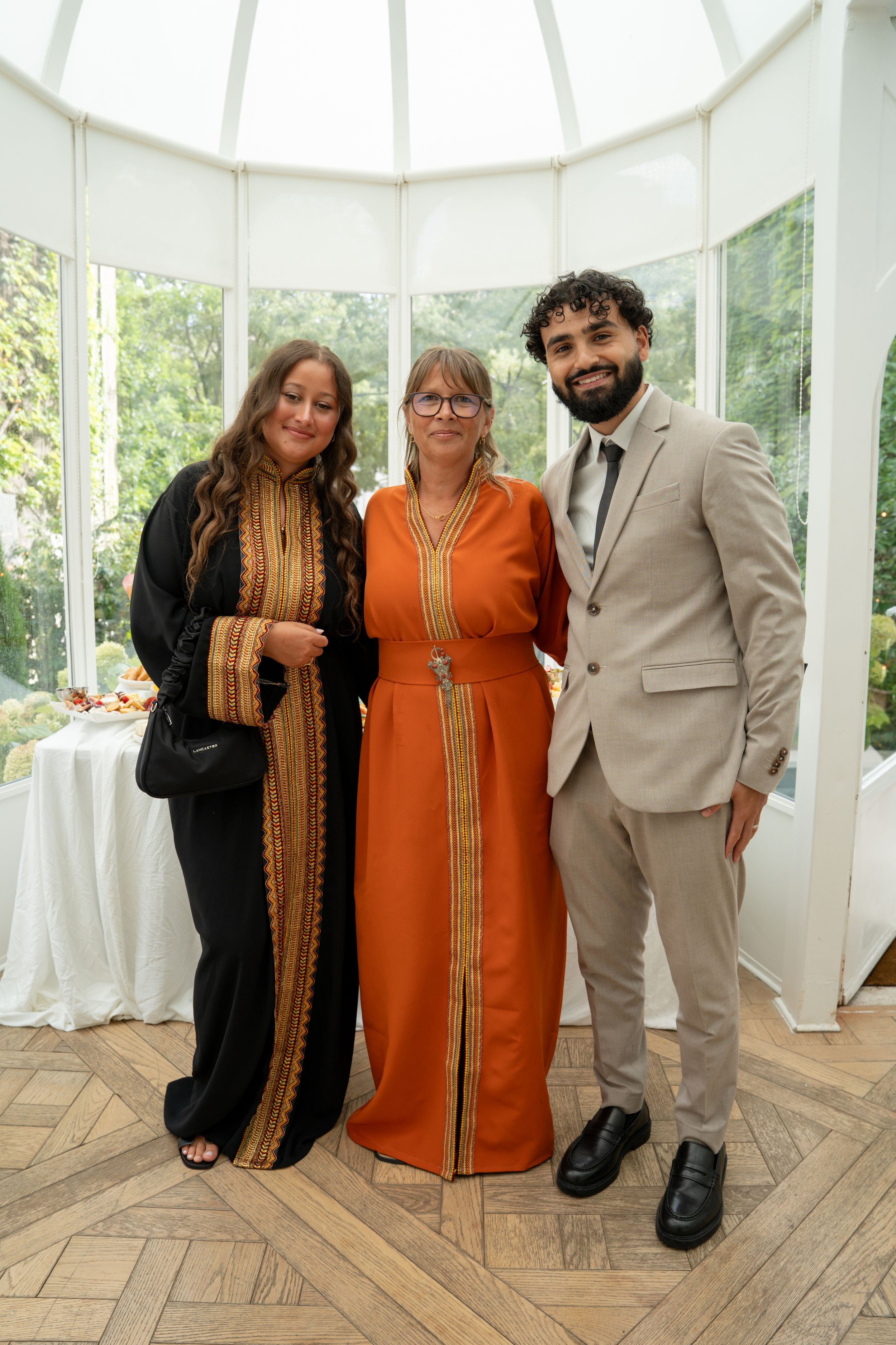 Three elegantly dressed people pose together in a bright conservatory at a formal event.