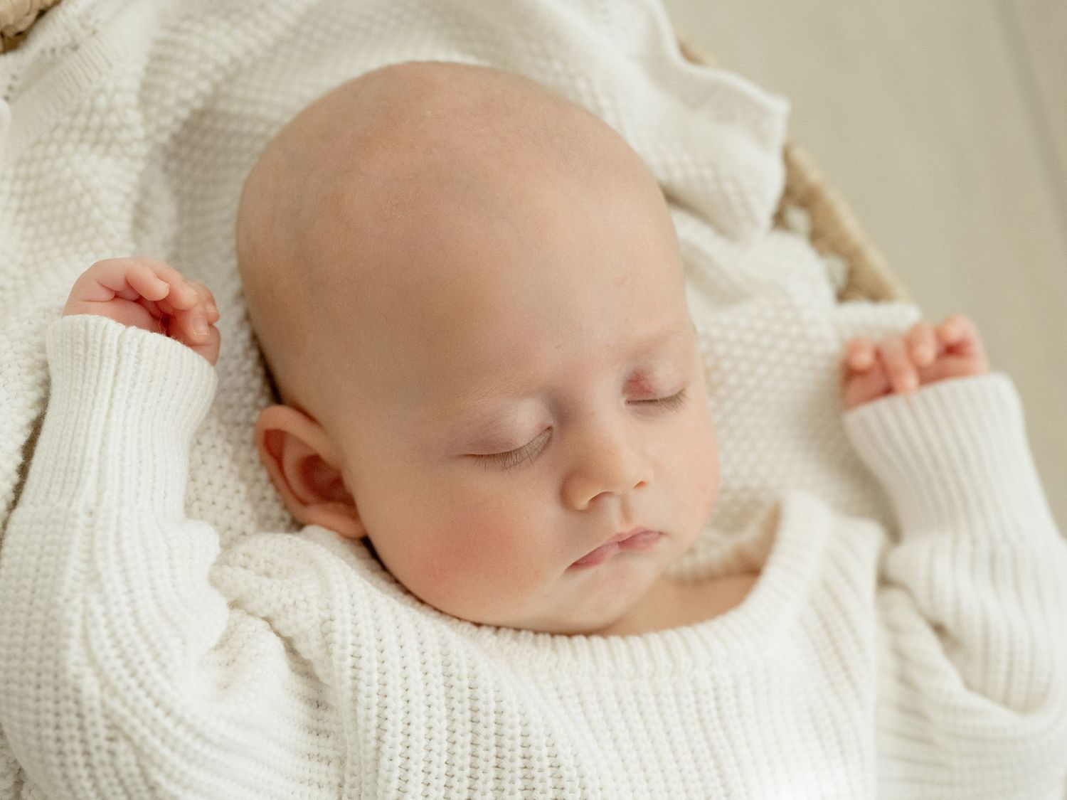 Peaceful newborn baby sleeping in a wicker basket, dressed in a white knit outfit with a soft white blanket.