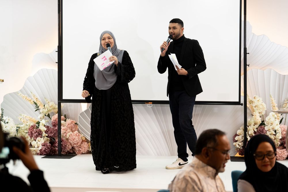 Two speakers present at a formal event with floral decorations and a projection screen backdrop.