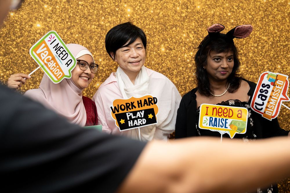 Three women pose with fun photo booth props including Work Hard Play Hard and I Need a Raise signs against a gold glitter backdrop.