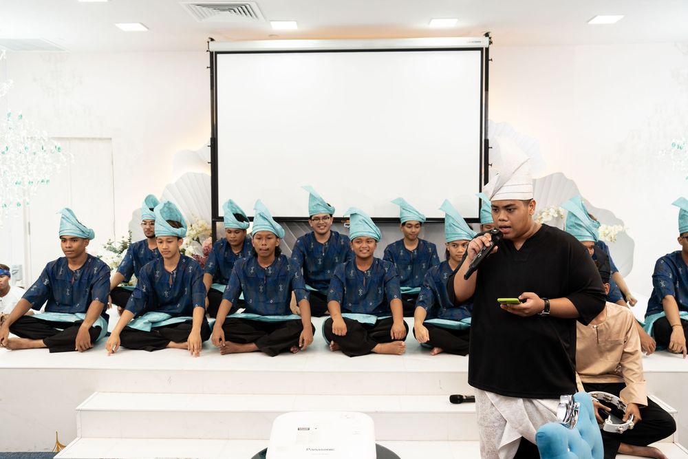 Group of students in blue uniforms and teal hats sit in rows during a classroom ceremony with a speaker presenting.