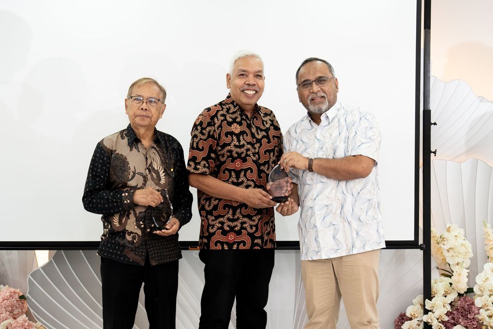 Three men in batik shirts smiling and posing together at a formal award ceremony indoors.