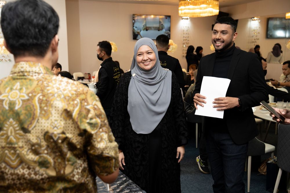 People in formal attire socializing at an indoor dining event, woman in hijab smiling at guests.