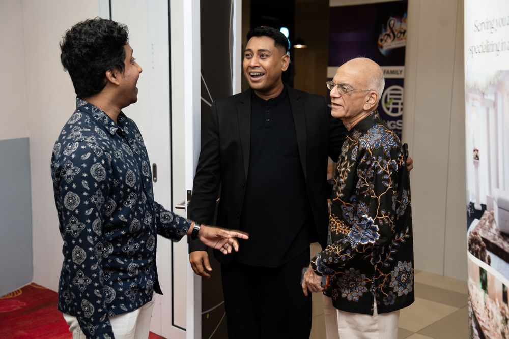 Three men in formal attire engaged in conversation at an indoor event, smiling warmly.