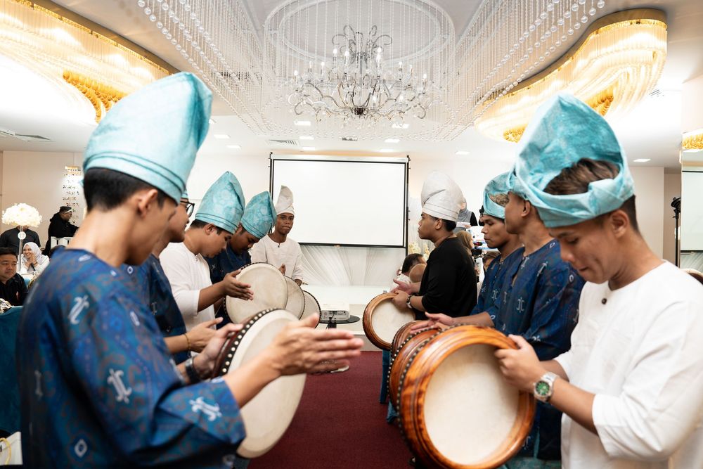 Young musicians in blue outfits and teal hats playing traditional drums at a cultural performance indoors.