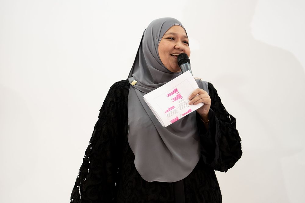 Woman in gray hijab and black cardigan speaks into microphone while holding papers at an event.