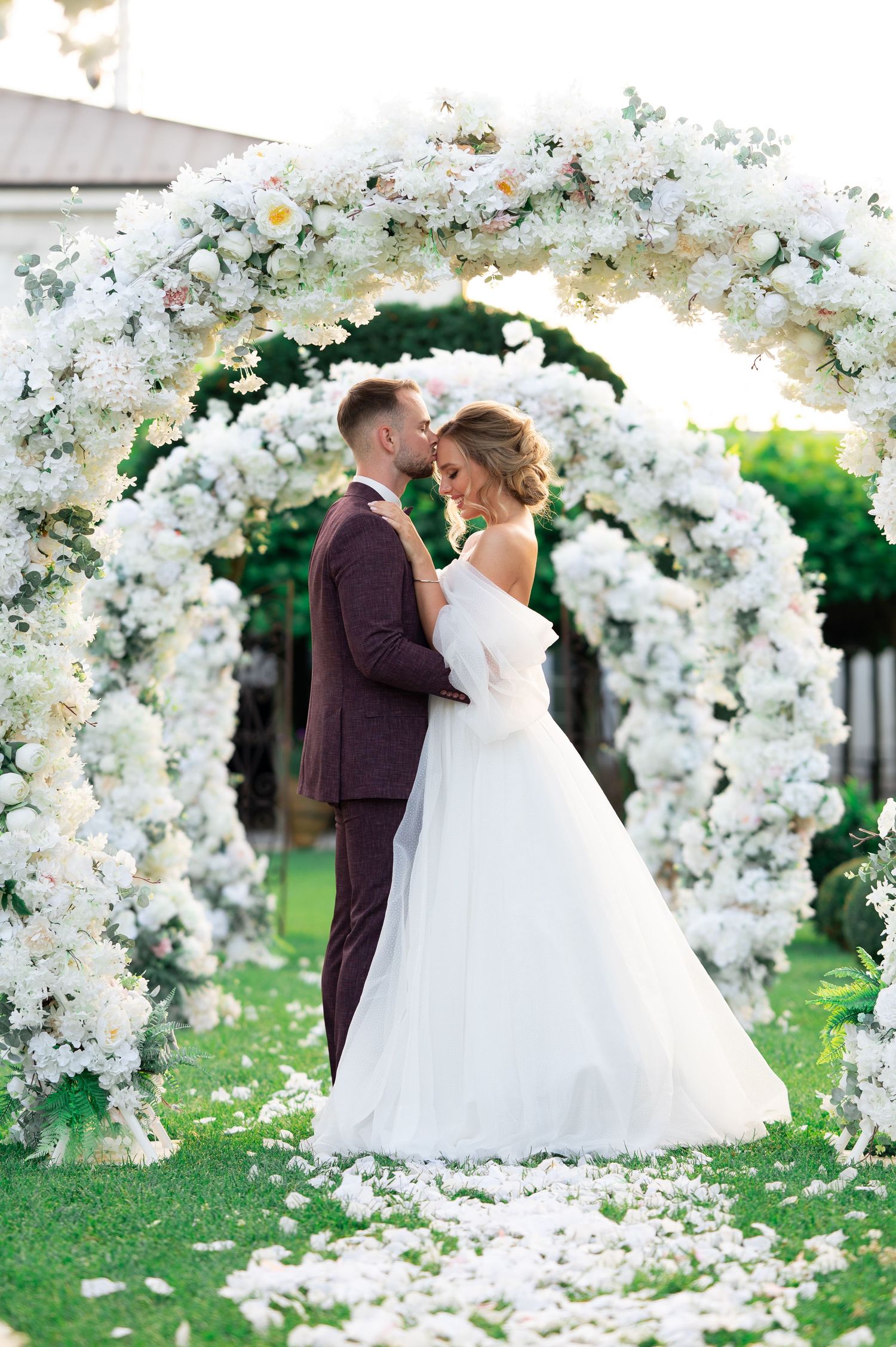 Couple shares romantic moment under stunning white floral arch on grass with rose petals.