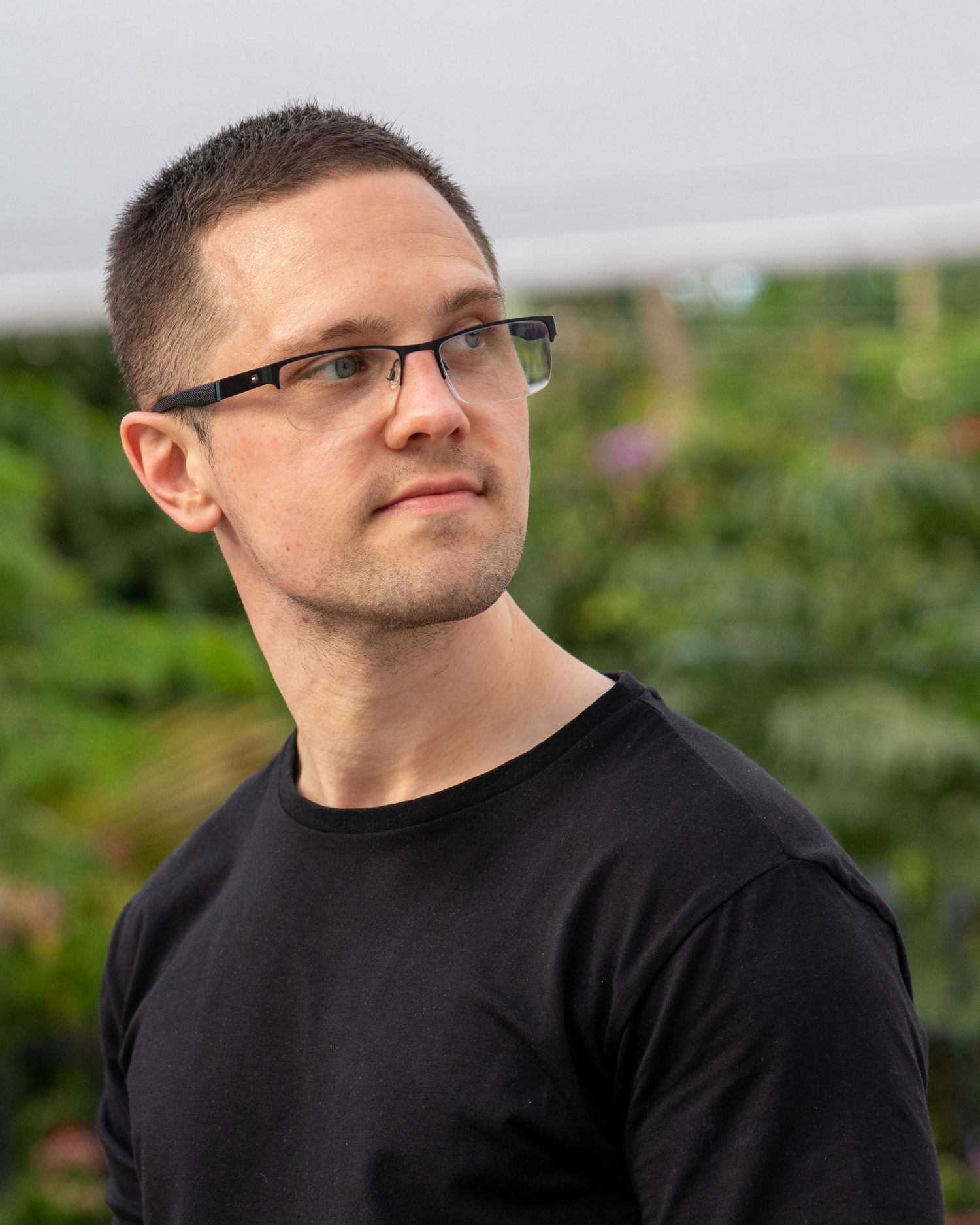 White man with short hair and glasses wearing black shirt poses outdoors with green foliage background.