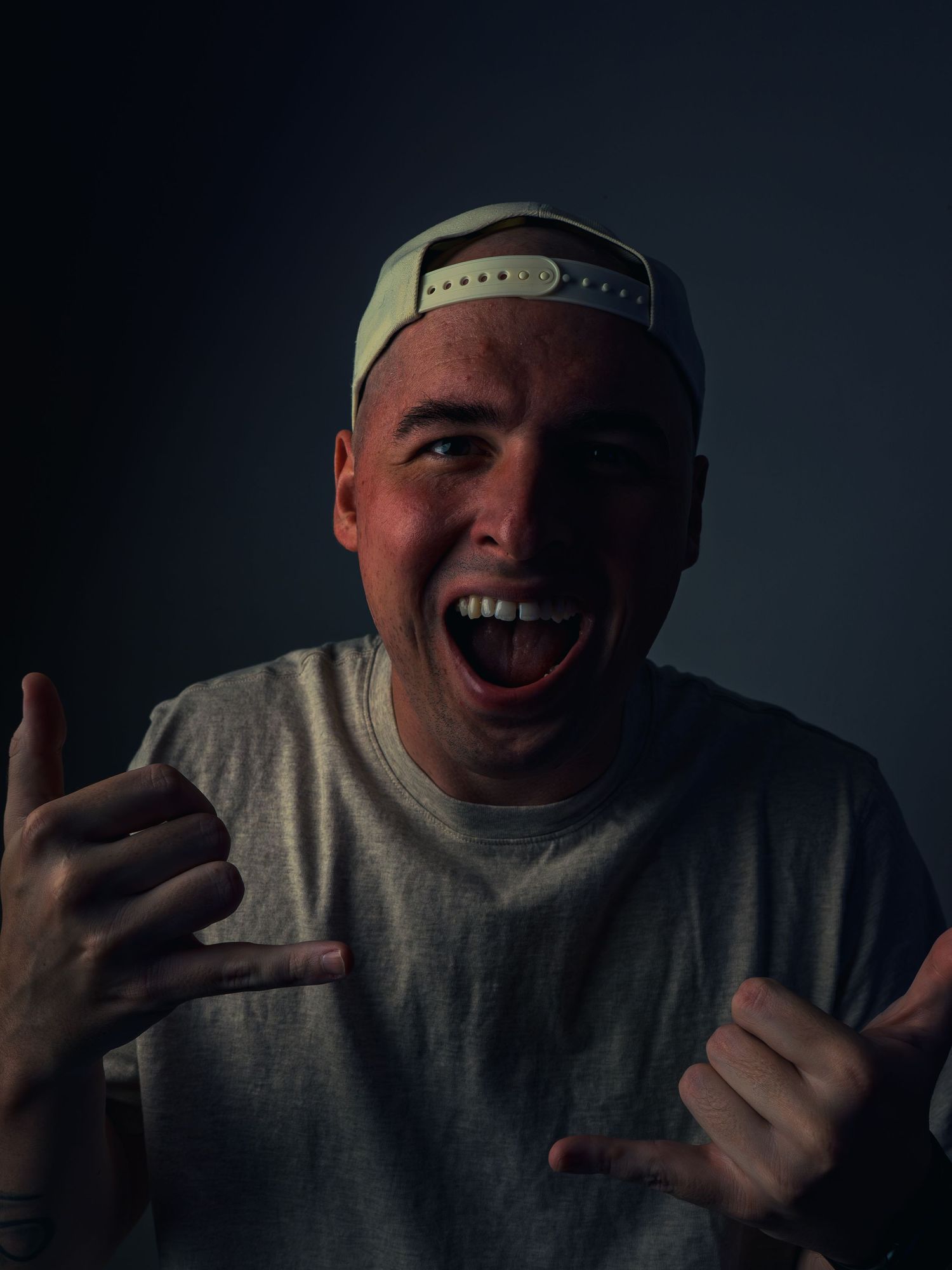 A man in a backward cap and gray tee laughs wide, throwing double shaka signs against a dark moody backdrop, a forearm tattoo showing