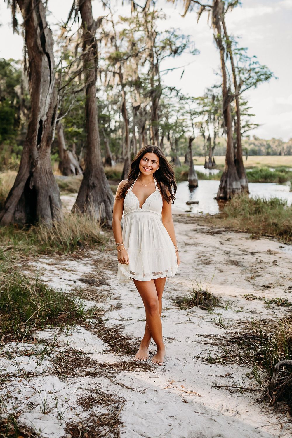 Young woman in white sundress standing barefoot on sandy path among cypress trees near water.