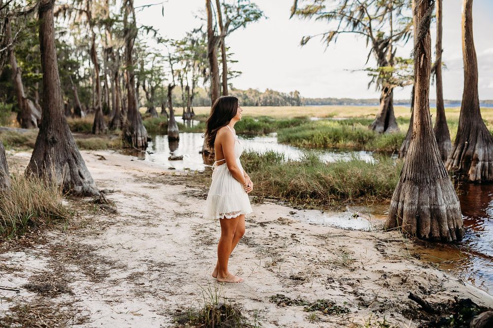 Young woman in white dress standing barefoot on sandy shore among cypress trees by a marsh at golden hour.