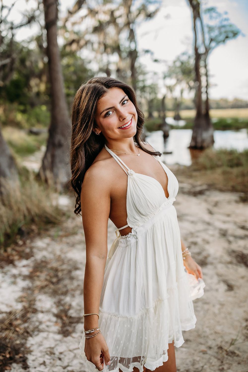 Young woman in white sundress smiling outdoors near a serene lake surrounded by trees.