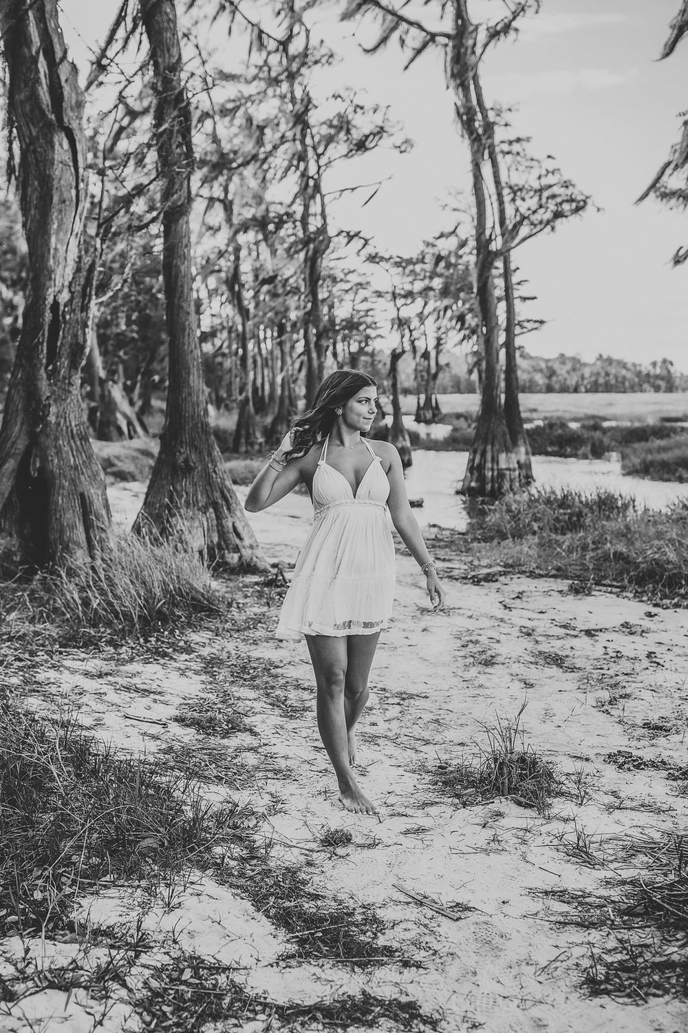 Black and white photo of a young woman in a white dress walking barefoot on sandy path among cypress trees near water.