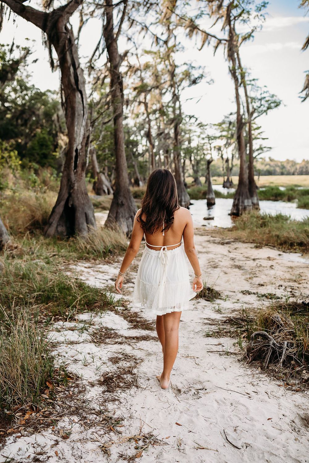 Woman in white dress walking barefoot on sandy path among cypress trees near water.