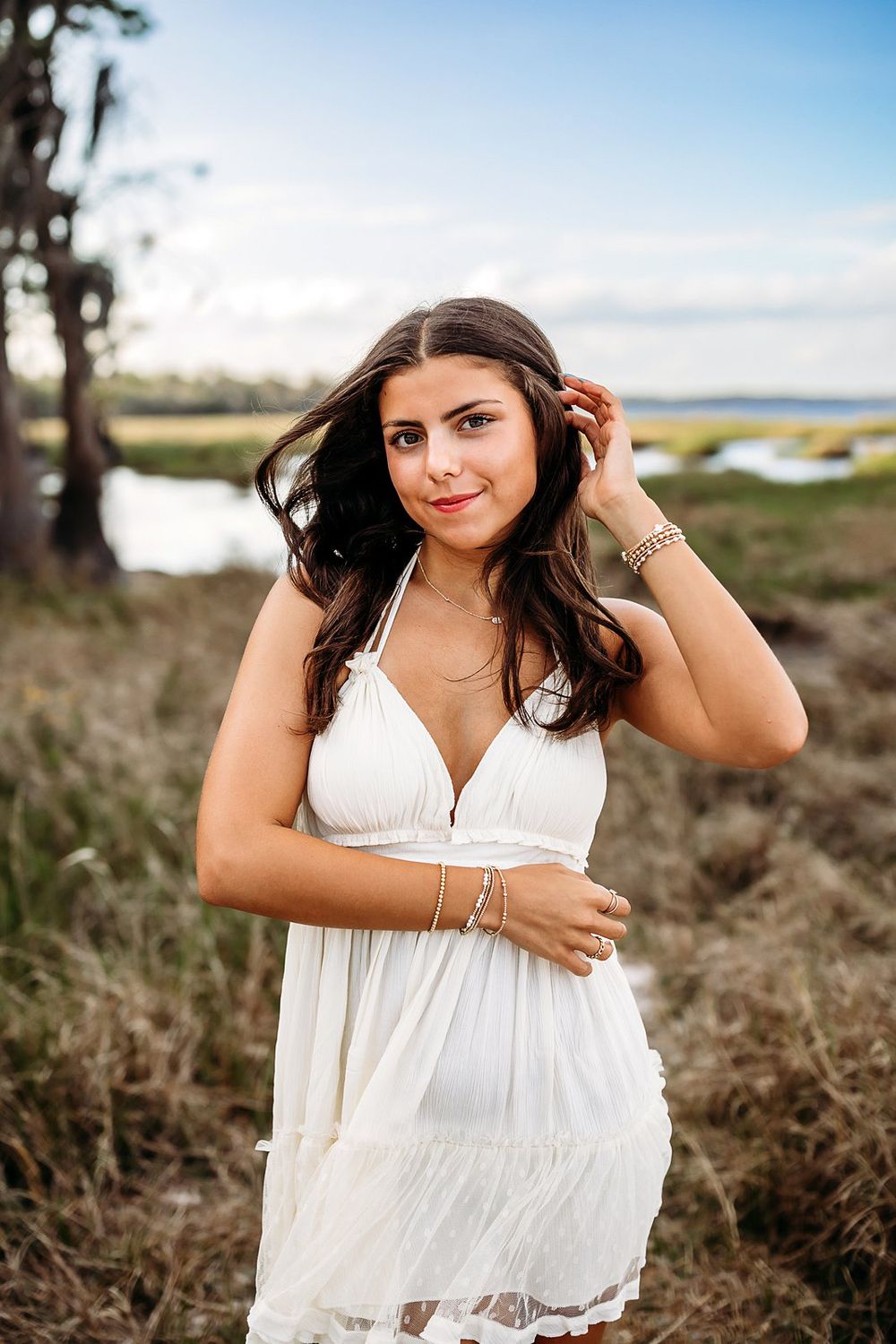 Young woman in white sundress poses outdoors near marsh, smiling with hand in dark hair on a sunny day.
