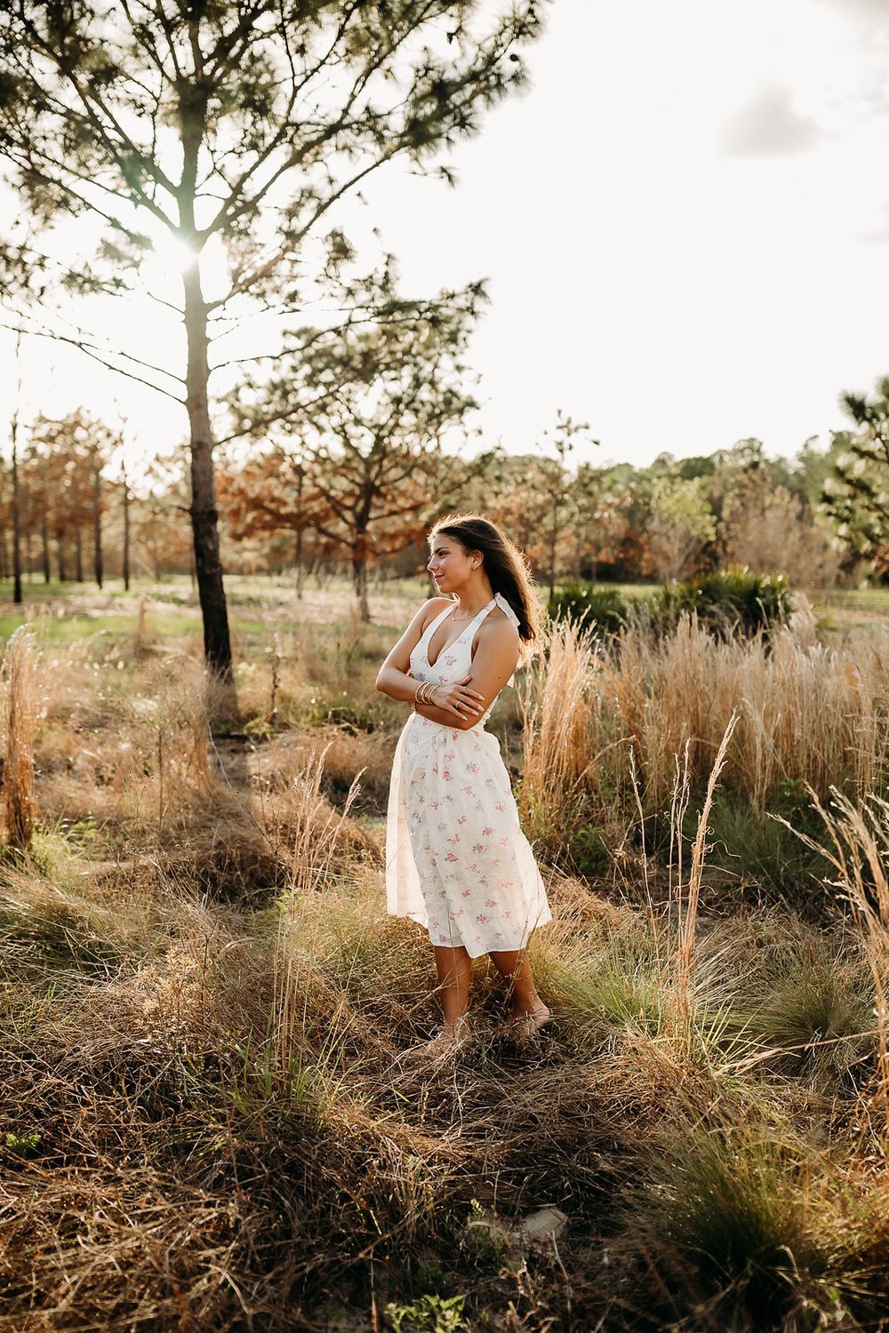 Woman in white floral midi dress standing in golden grass field surrounded by trees during sunset.