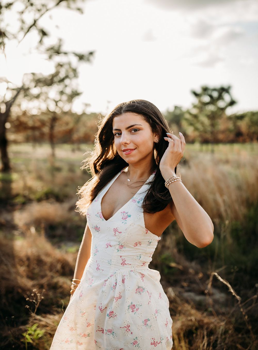 Young woman in floral sundress poses outdoors in golden hour light among natural grasslands.