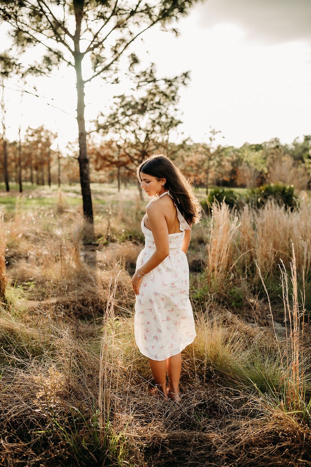 Young woman in white floral dress standing barefoot in golden grass field at sunset.