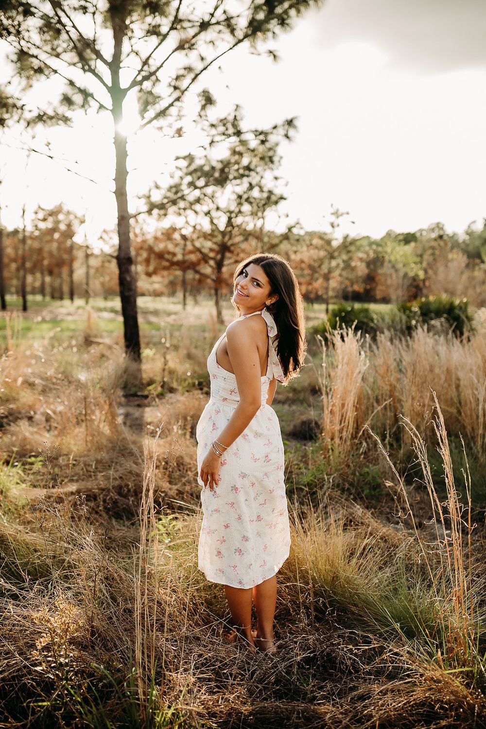 Young woman in floral sundress standing in golden field at sunset, looking over shoulder.