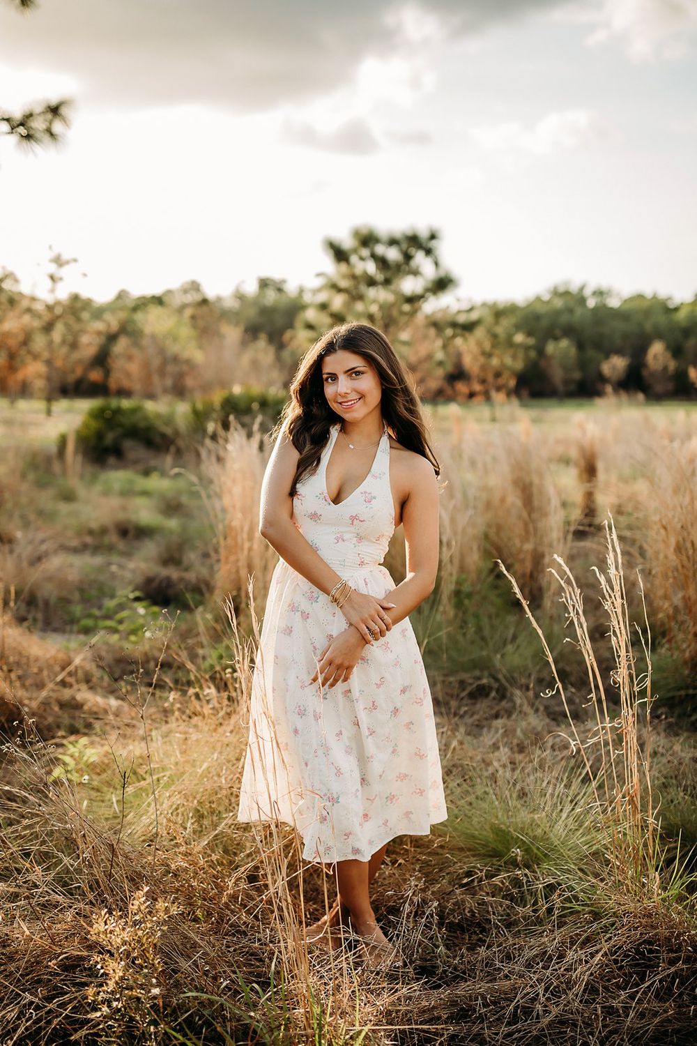 Young woman in floral midi dress standing in golden field at sunset, smiling outdoors.