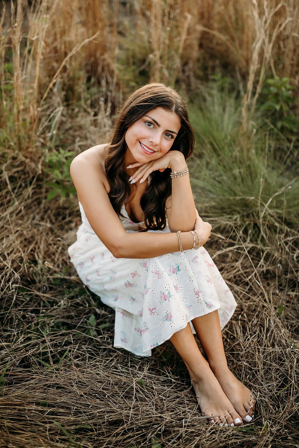 Young woman in white floral dress sitting barefoot in a golden grass field, smiling warmly at the camera.