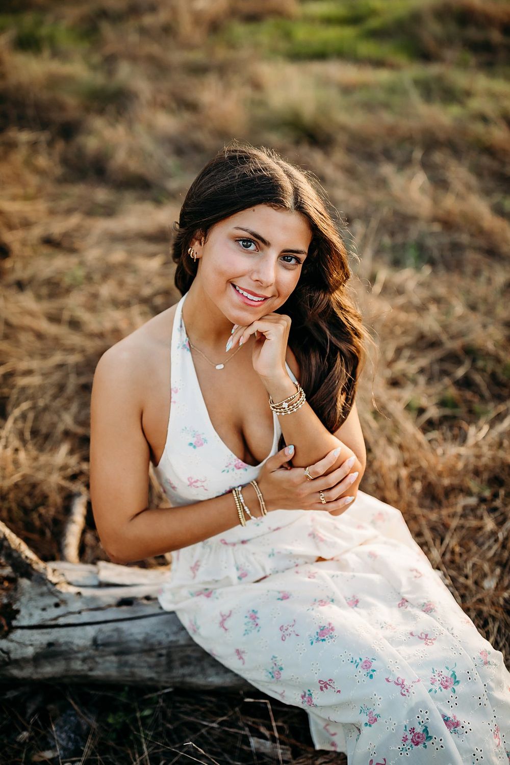 Young woman in floral sundress sitting on log in golden field, smiling warmly at camera.