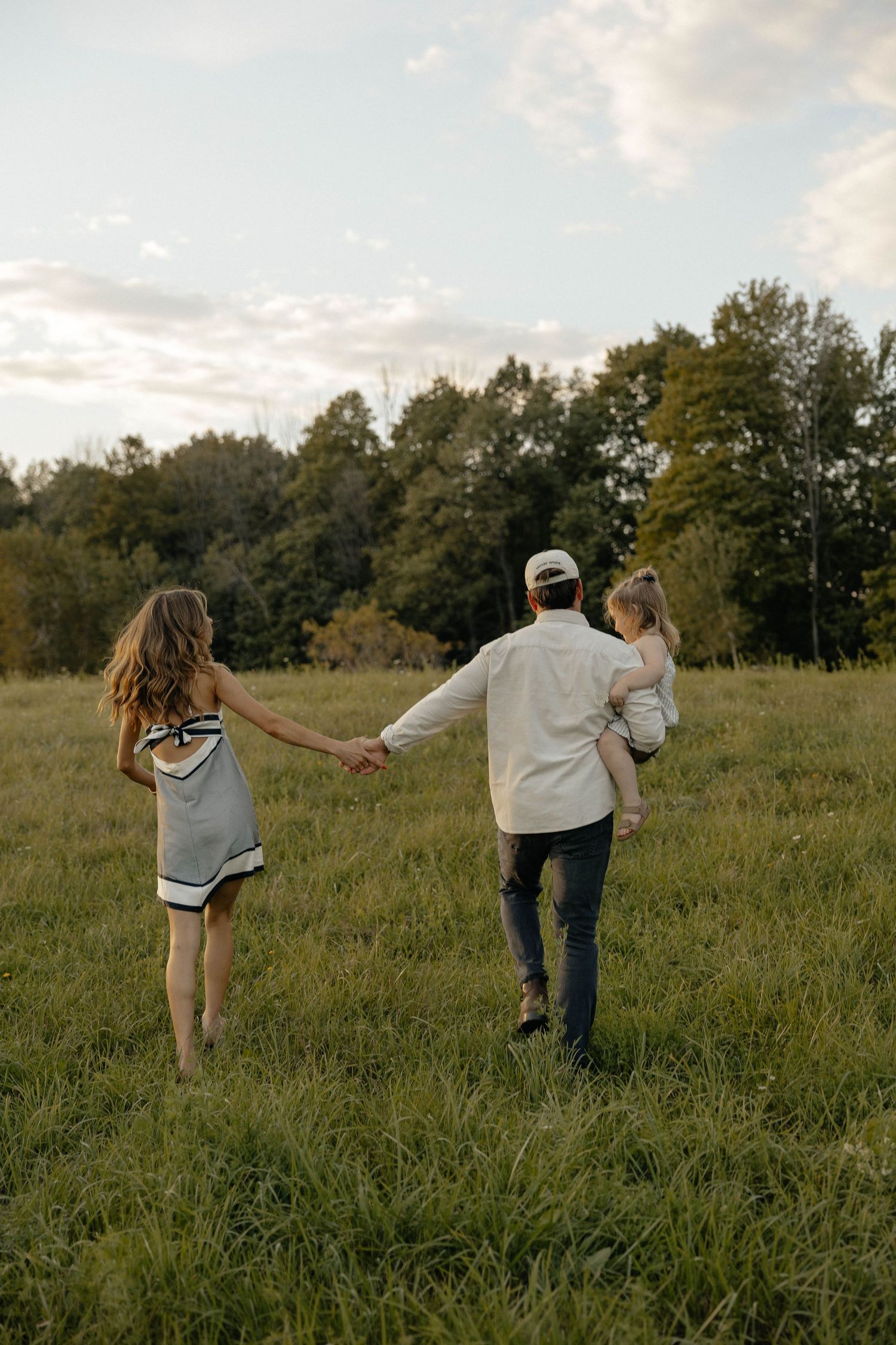 A family of three walks through a green meadow, parents holding hands with dad carrying their young child.