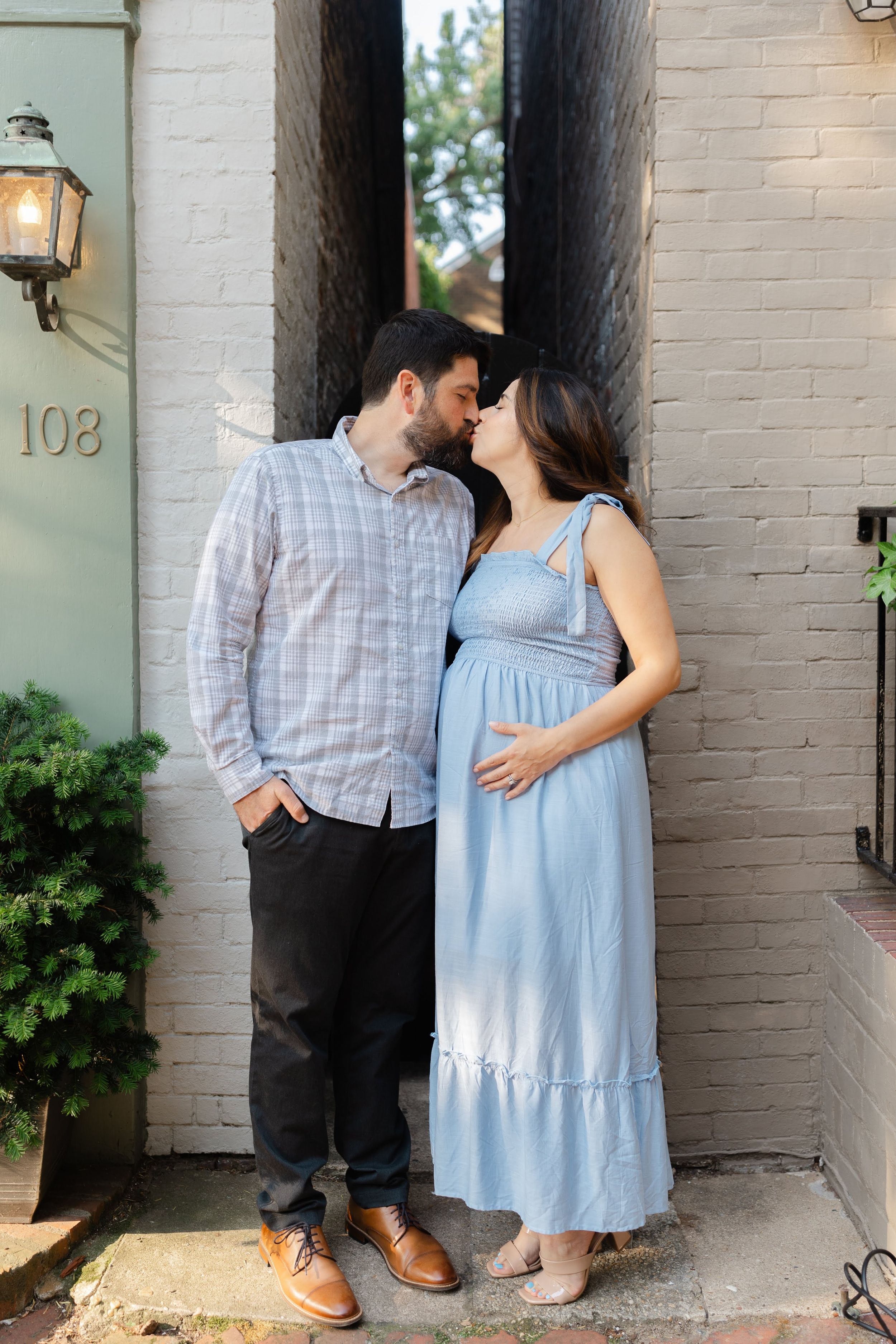 Pregnant couple sharing a kiss outside a brick building, woman in blue maxi dress cradling baby bump.