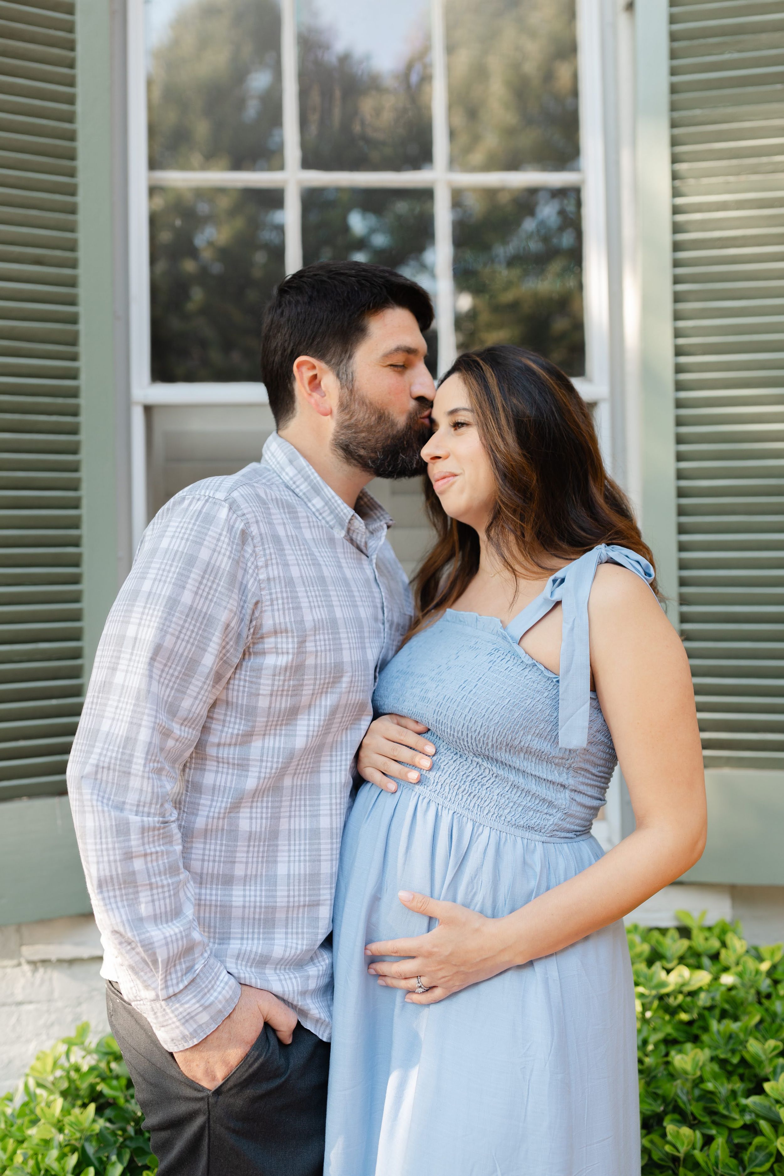 A man kisses a pregnant woman's forehead as she smiles, both dressed in blue, standing outside a home.